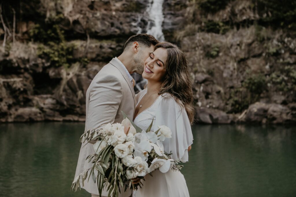 groom kissing bride on cheek in front of waterfall Hawaii adventure wedding