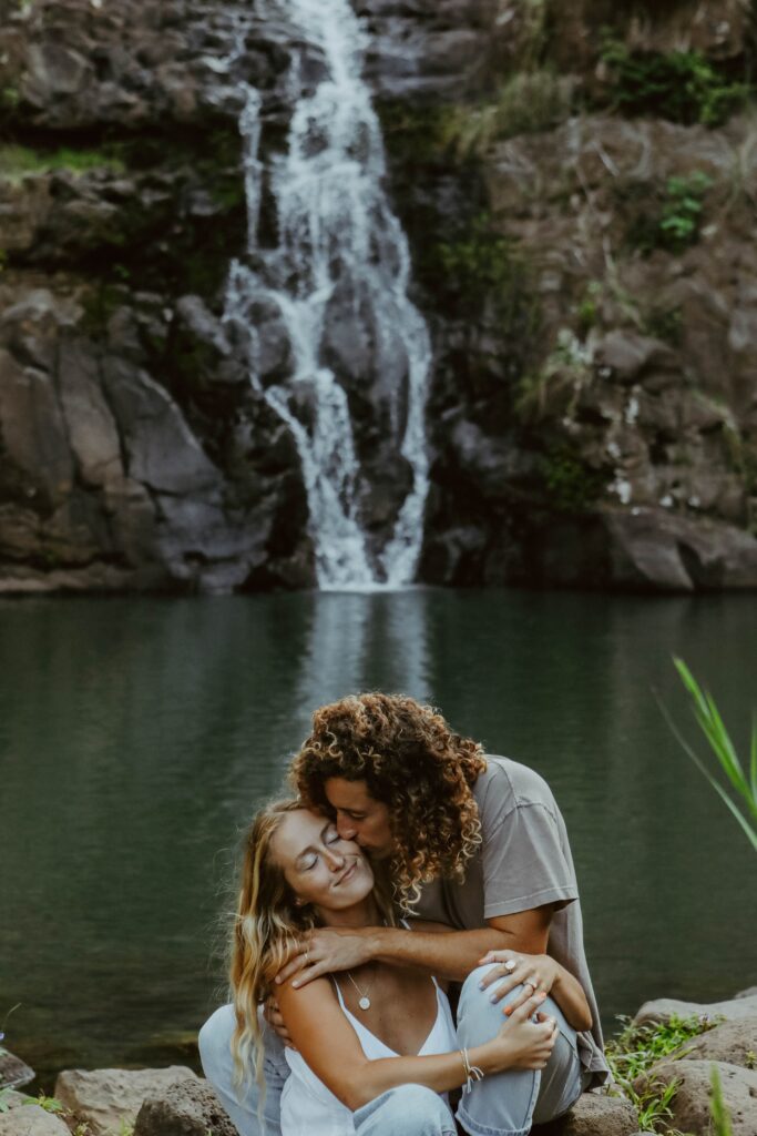 husband kissing wife on cheek in front of waterfall in Hawaii Elopement packages near me 